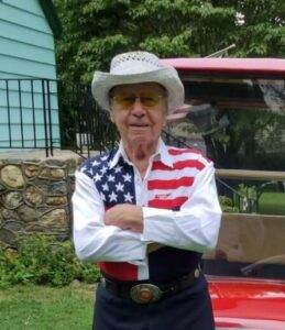 1. Elderly man wearing patriotic outfit and cowboy hat outdoors in front of a red vehicle.
