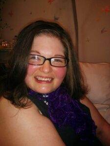 Warm smiling woman with glasses, dark hair, and purple scarf, relaxed and happy.