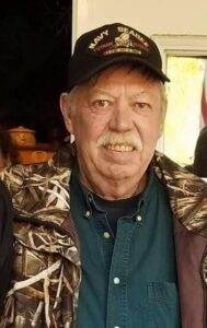 Gentleman wearing a navy cap and camouflage jacket, smiling indoors, in a warm, welcoming setting.